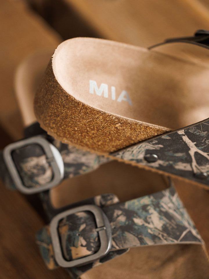 A close-up of The Kane by Savannah Sevens western life{&}style, a camo slip-on sandal with a cork sole, camouflage straps, and metal buckles. The sandal is displayed on a wooden surface.