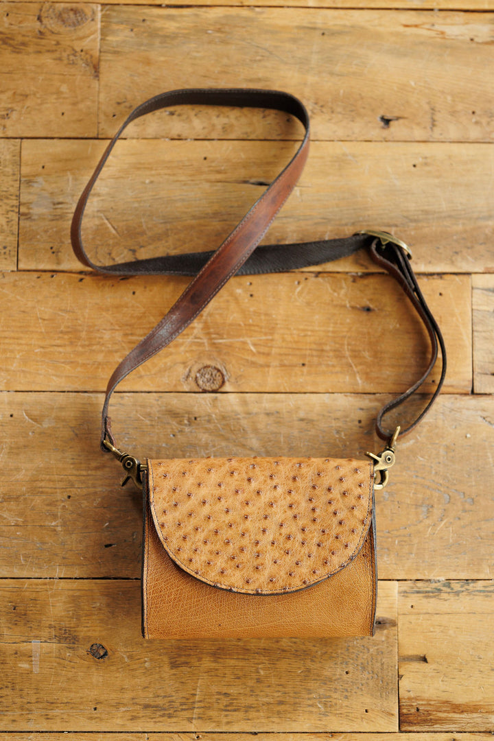 The Chicota by Savannah Sevens western life{&}style, a textured tan ostrich leather crossbody with a dark brown strap, rests on a rustic wooden surface.