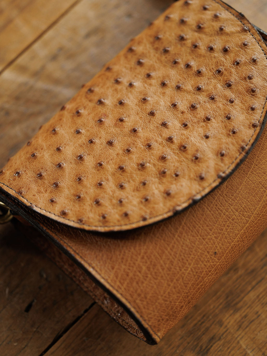 A close-up of The Chicota by Savannah Sevens western life{&}style, a saddle tan ostrich leather clutch with a textured pattern, elegantly displayed on a wooden surface.