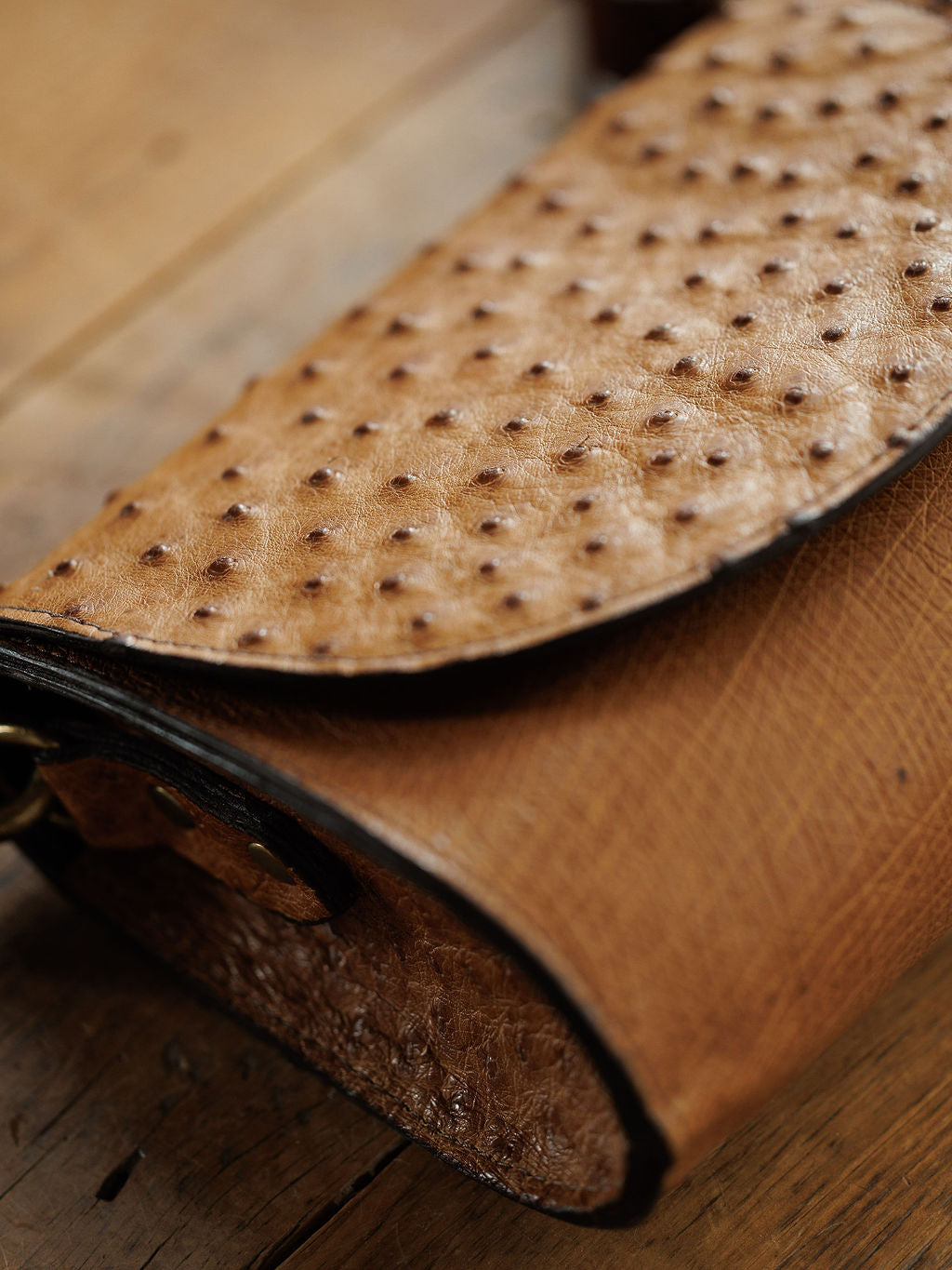 A close-up of The Chicota by Savannah Sevens western life{&}style, a saddle tan ostrich leather clutch with raised bumps and a curved flap, resting on a wooden surface.