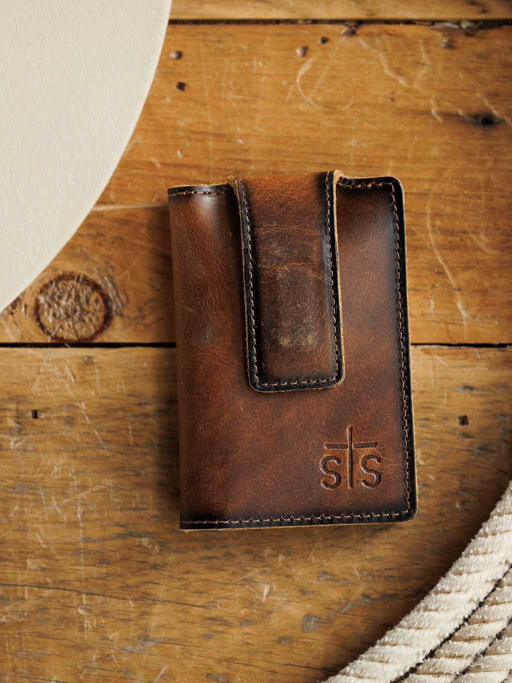 The Waynesboro Money Clip Wallet by Savannah Sevens western life{&}style, a brown full grain leather wallet with visible stitching and an embossed sts logo, sits on rustic wood near a white round object and coiled rope.