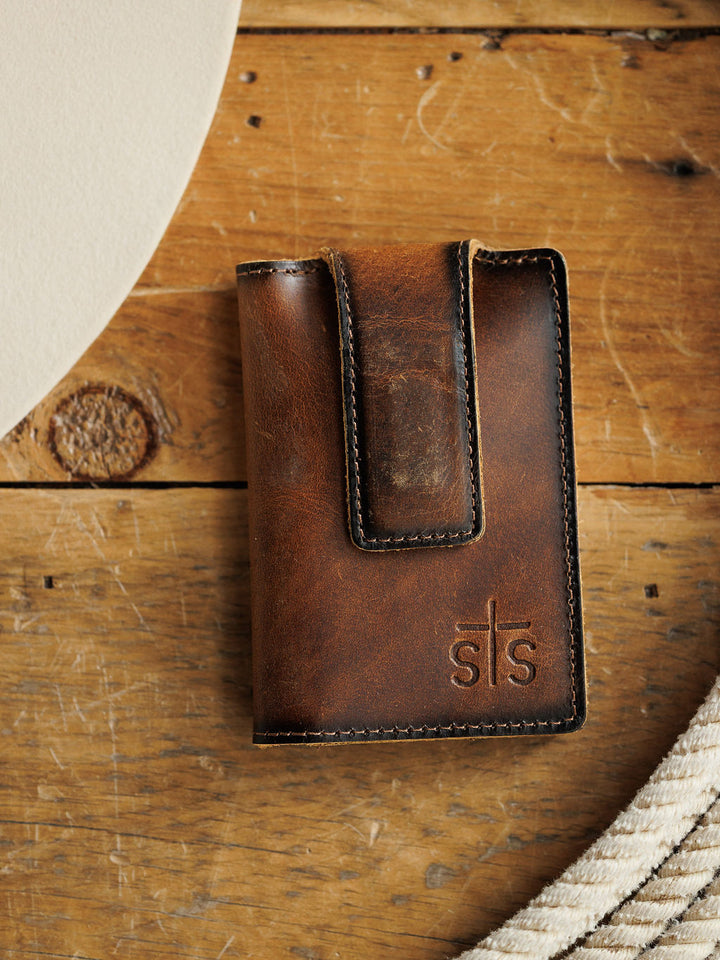 The Waynesboro Money Clip Wallet by Savannah Sevens western life{&}style, a brown full grain leather wallet with visible stitching and an embossed sts logo, sits on rustic wood near a white round object and coiled rope.