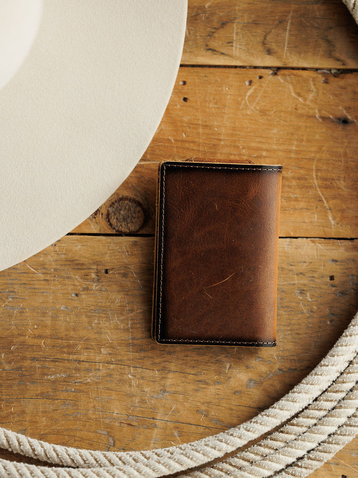 The Waynesboro Money Clip Wallet by Savannah Sevens western life{&}style, crafted from brown full grain leather, rests on a rustic wooden surface beside a coiled white rope and the edge of a light-colored hat.