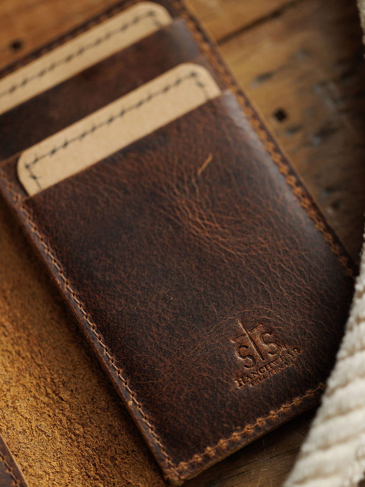 A close-up of the Waynesboro Money Clip Wallet by Savannah Sevens western life{&}style, crafted from full grain leather with two card slots holding tan cards, and embossed branding near the bottom edge on a wooden surface.