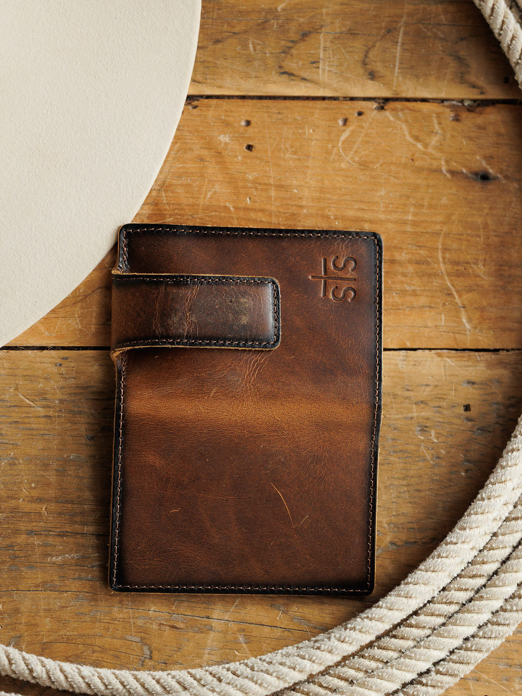 The Waynesboro Money Clip Wallet by Savannah Sevens western life{&}style, crafted from full grain leather with visible stitching and an embossed logo, rests on a rustic wooden surface beside a white rope and a round beige object.