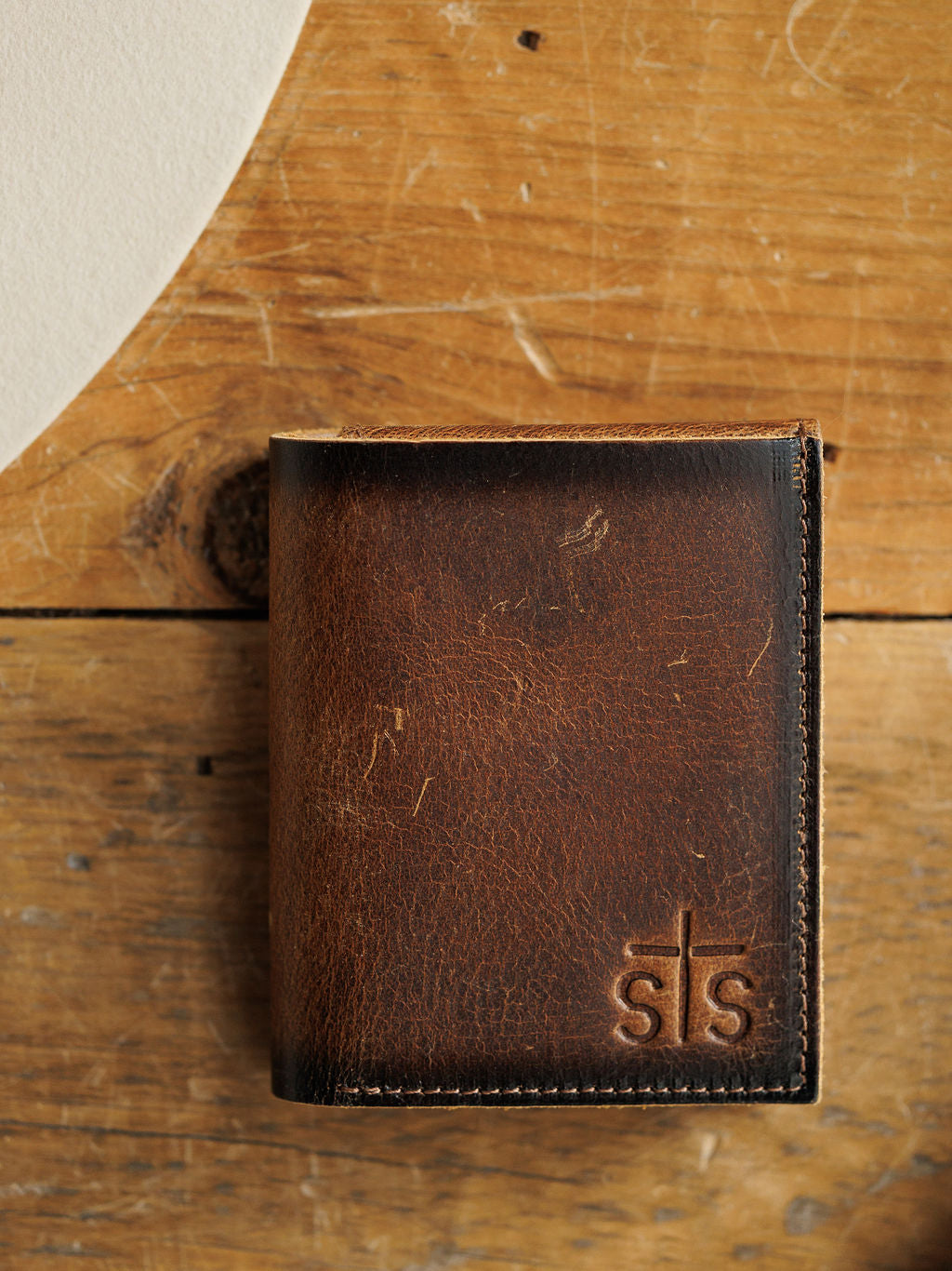The Waynesboro Hidden Cash Wallet by Savannah Sevens western life{&}style, a brown distressed full grain leather men's wallet with an embossed sts logo, is displayed on a rustic wooden surface.