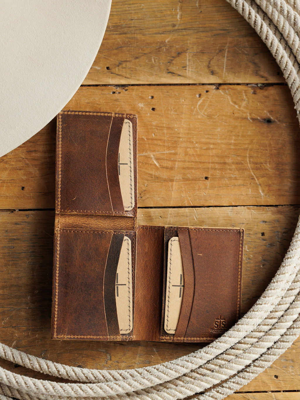 The Waynesboro Hidden Cash Wallet by Savannah Sevens western life{&}style, made of distressed full grain leather with tan card slots, is displayed open on a rustic wooden surface beside a coiled rope and a round white object.
