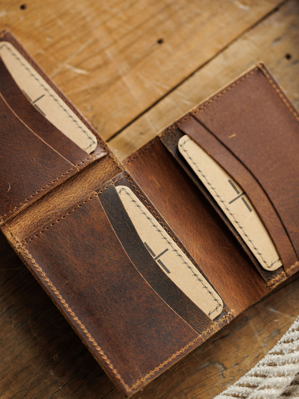 The Waynesboro Hidden Cash Wallet by Savannah Sevens western life{&}style, crafted from distressed full grain leather, lies open on a wooden surface displaying multiple card slots and tan stitching that highlight its rugged texture and craftsmanship.