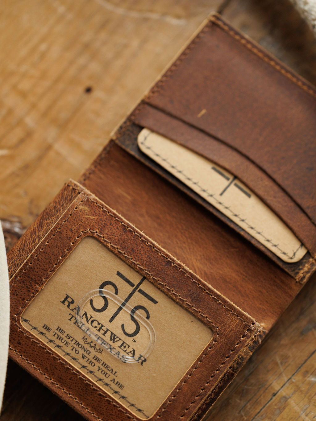 The Waynesboro Hidden Cash Wallet by Savannah Sevens western life{&}style is a brown distressed full grain leather men’s wallet with visible stitching, clear ID slot, and card slot, shown resting on a wooden surface.
