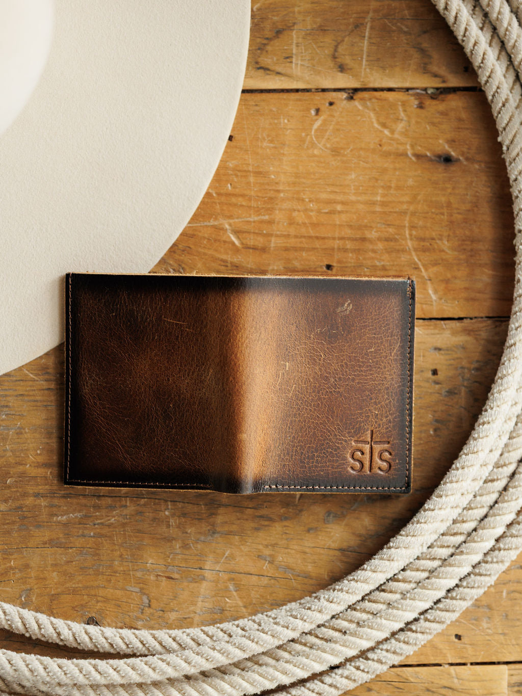 The Waynesboro Hidden Cash Wallet by Savannah Sevens western life{&}style, crafted from distressed full grain leather with “sTs” embossed in the corner, rests on a wooden surface beside a coiled rope and a light-colored hat.