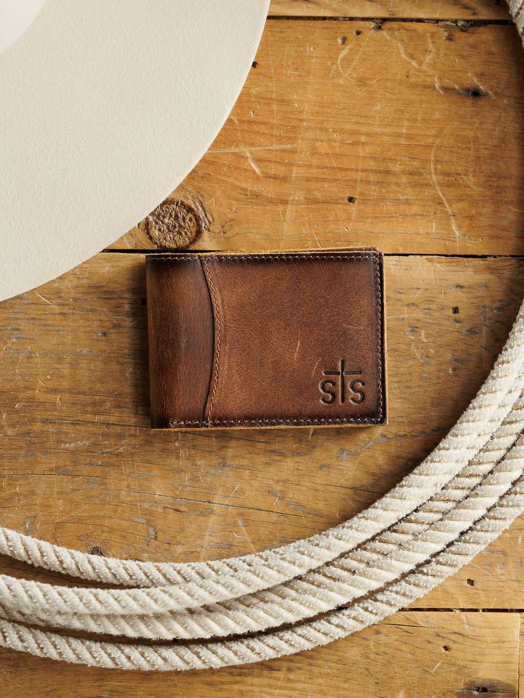 The Waynesboro Bifold Wallet by Savannah Sevens western life{&}style features distressed full grain leather with a cross embossed on the corner, shown on wood next to a white cowboy hat and coiled rope.