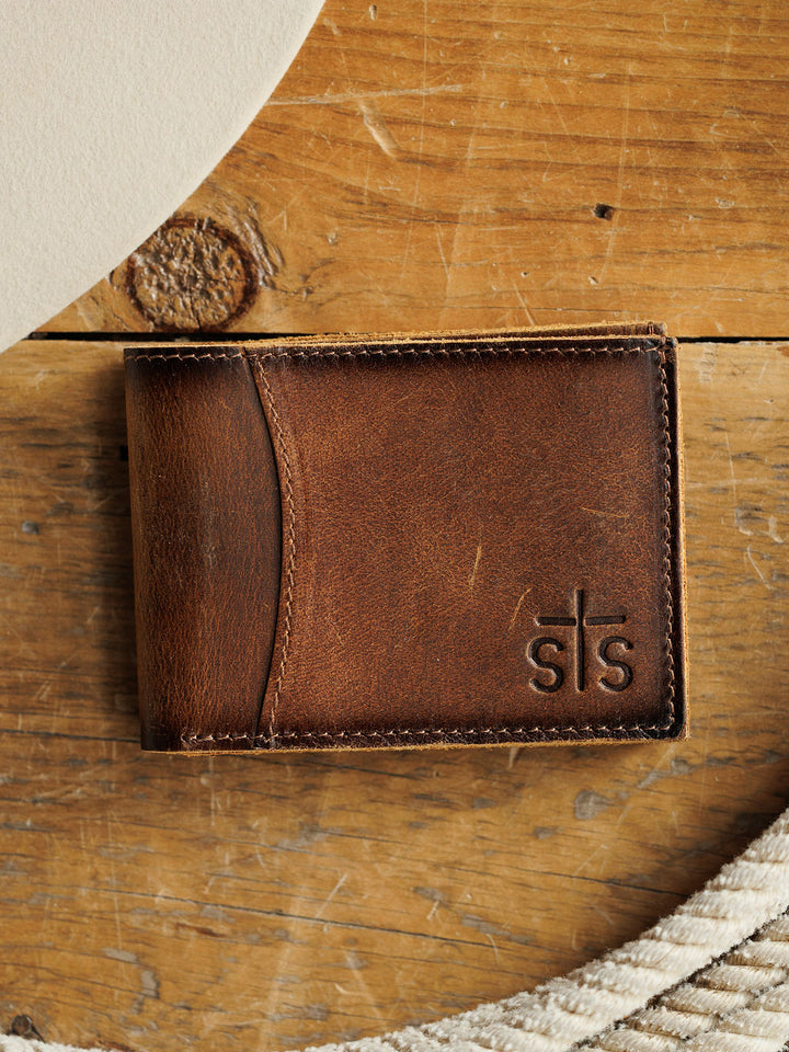 The Waynesboro Bifold Wallet by Savannah Sevens western life{&}style is a men's distressed full grain leather wallet with visible stitching and "STS Ranch" embossed on the corner, shown on a wooden surface next to cream circular fabric.