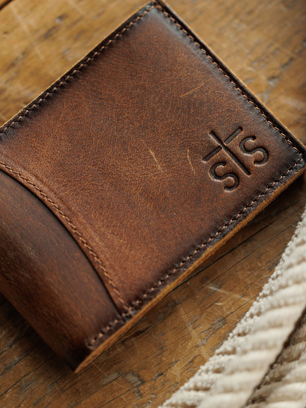 A close-up of a Savannah Sevens western lifestyle Waynesboro Bifold Wallet, crafted from distressed full grain leather with embossed initials, resting on a wooden surface next to a coiled rope.