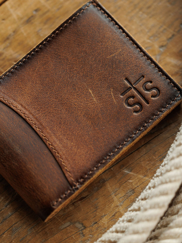 A close-up of a Savannah Sevens western lifestyle Waynesboro Bifold Wallet, crafted from distressed full grain leather with embossed initials, resting on a wooden surface next to a coiled rope.