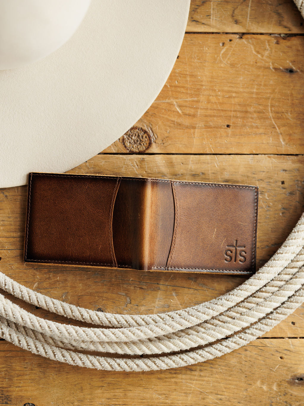 A Savannah Sevens western life{&}style Waynesboro Bifold Wallet made from distressed full grain leather sits on a wooden surface next to a white cowboy hat and coiled rope.