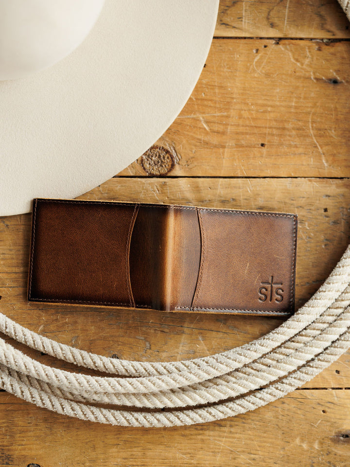 A Savannah Sevens western life{&}style Waynesboro Bifold Wallet made from distressed full grain leather sits on a wooden surface next to a white cowboy hat and coiled rope.