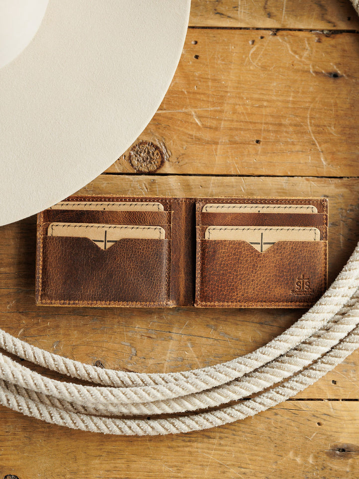 The Waynesboro Bifold Wallet by Savannah Sevens western life{&}style, crafted from brown distressed full grain leather, lies open on a rustic wooden surface with tan cards inside. A white hat and coiled rope sit nearby.