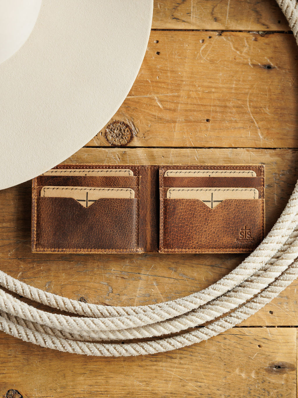 The Waynesboro Bifold Wallet by Savannah Sevens western life{&}style is a brown men's wallet crafted from distressed full grain leather, shown open with four card slots on a rustic wood surface beside a white cowboy hat and coiled rope. Inspired by STS Ranch style.
