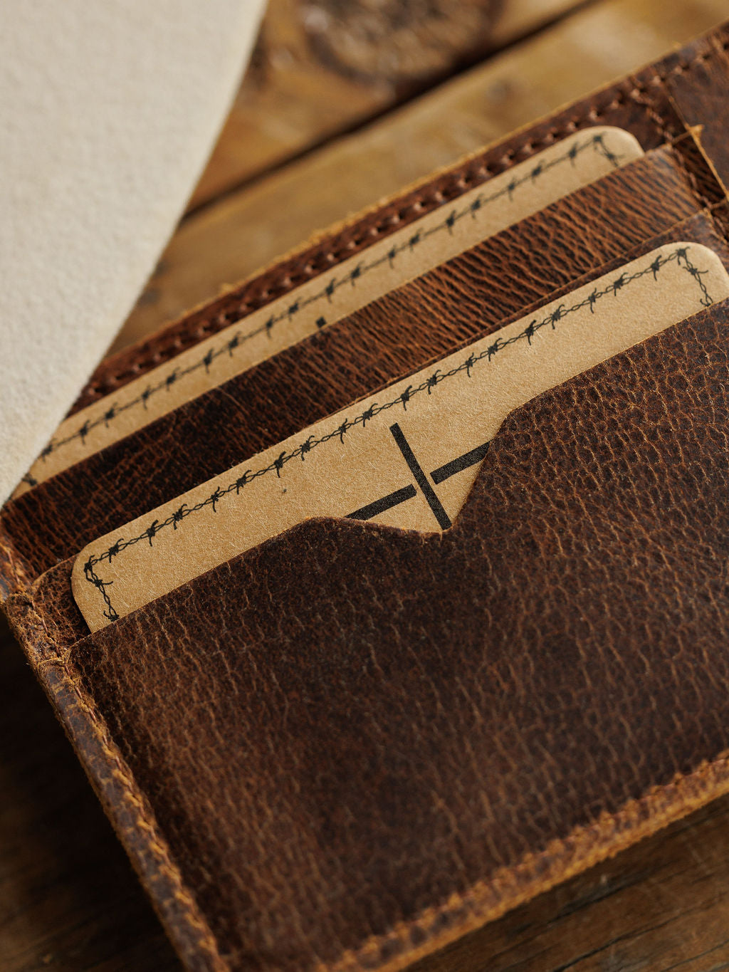 The Waynesboro Bifold Wallet by Savannah Sevens western life{&}style is a men's distressed full grain leather bifold, shown on wood holding two brown cards with black stitching accents.