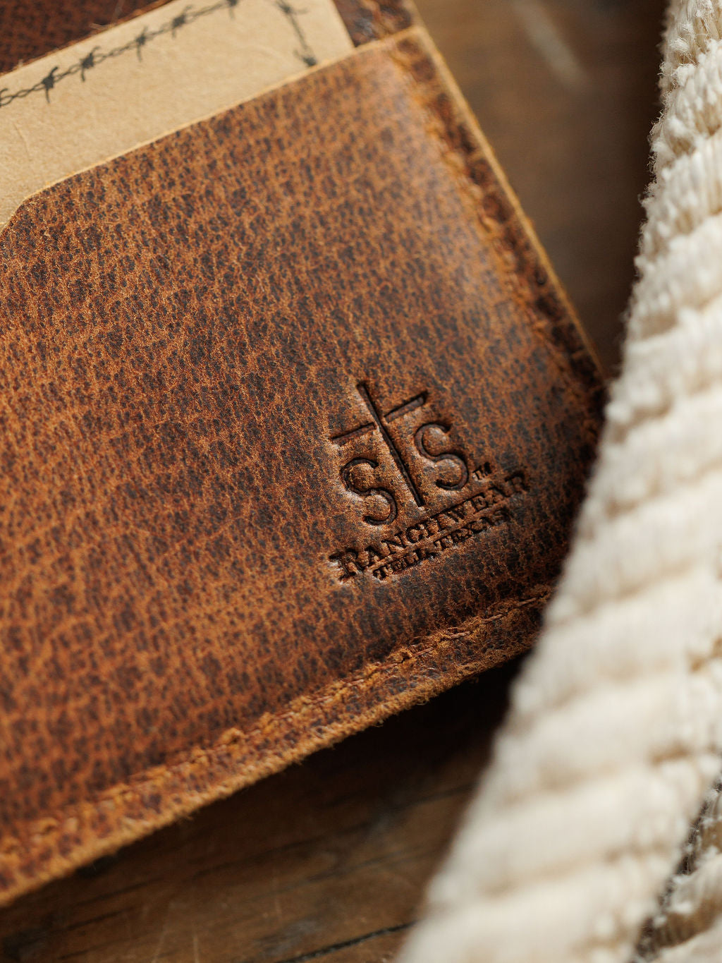 Close-up of the Waynesboro Bifold Wallet by Savannah Sevens western life{&}style, featuring distressed full grain leather, visible stitching, and an embossed logo, resting on textured cream-colored fabric.