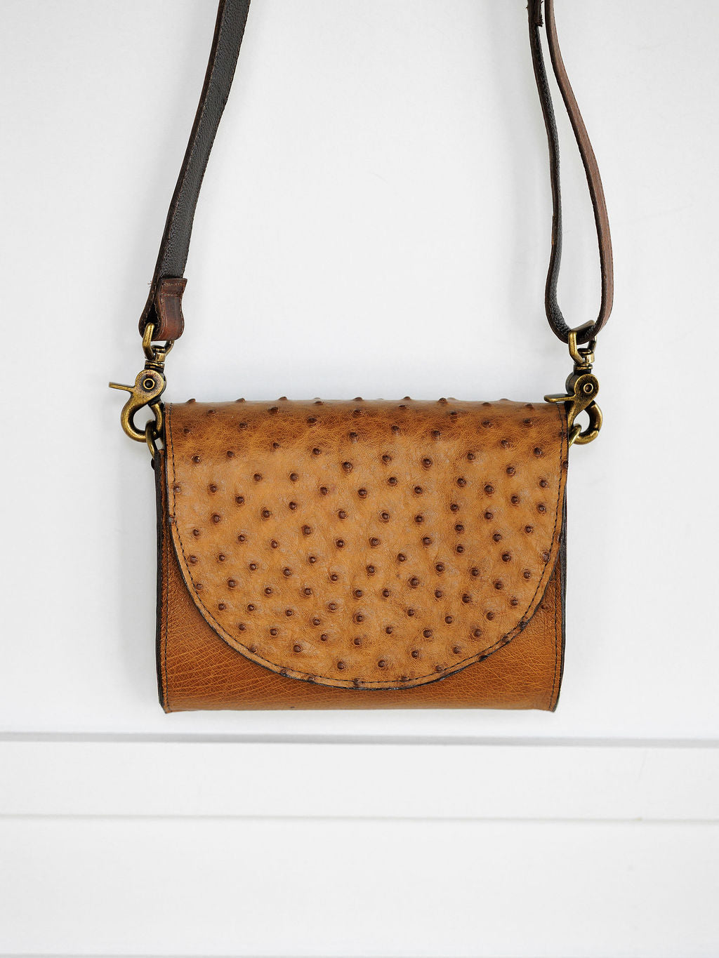 The Chicota by Savannah Sevens western life{&}style: a small women's clutch in saddle tan ostrich leather with a textured, spotted flap and long adjustable strap, shown against a white background.