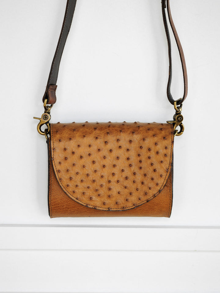 The Chicota by Savannah Sevens western life{&}style: a small women's clutch in saddle tan ostrich leather with a textured, spotted flap and long adjustable strap, shown against a white background.