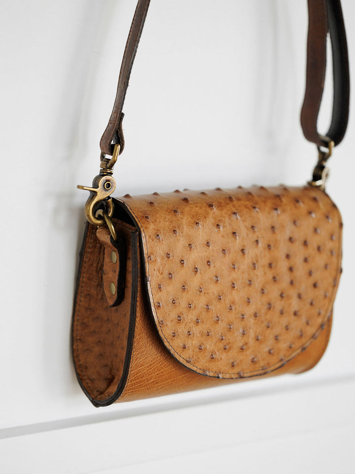 The Chicota by Savannah Sevens western life{&}style is a brown ostrich leather crossbody with a dimpled texture, brass hardware, flap closure, and dark brown adjustable strap, shown hanging on a white wall.