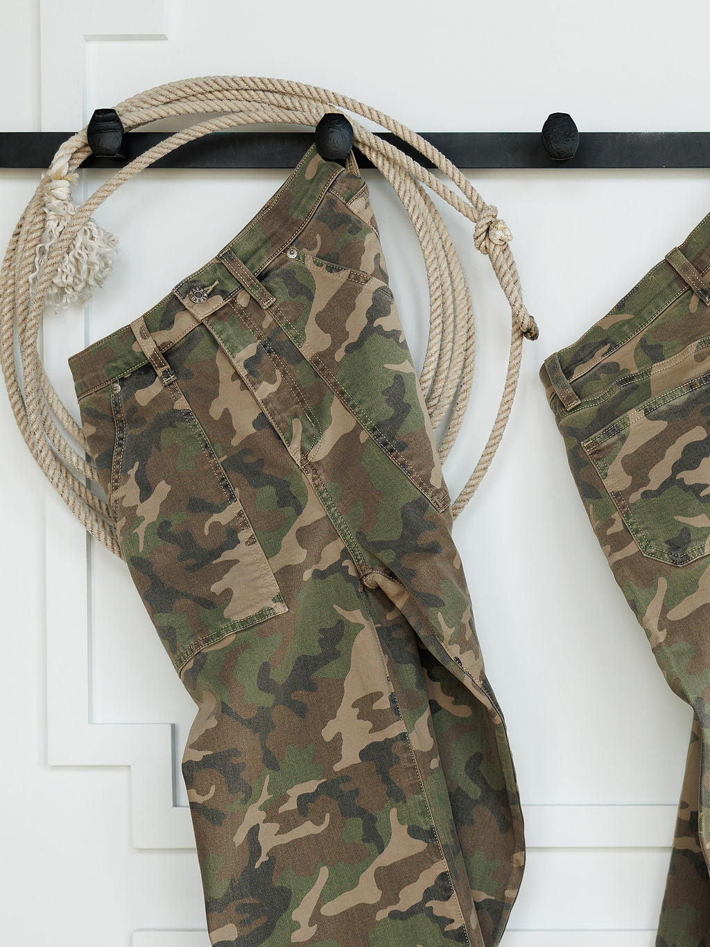 The Granger High Rise Wide Leg Jean by Savannah Sevens western life{&}style, featuring a camo pattern, hangs on a black wall hook with a coiled rope beside it against a white background.