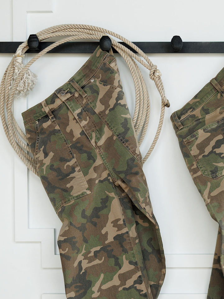 The Granger High Rise Wide Leg Jean by Savannah Sevens western life{&}style, featuring a camo pattern, hangs on a black wall hook with a coiled rope beside it against a white background.