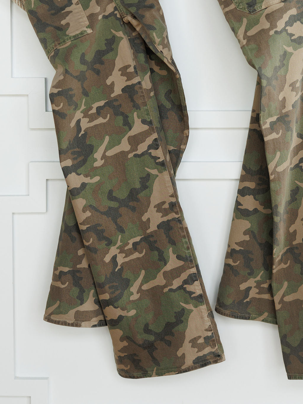Close-up of two pairs of The Granger High Rise Wide Leg Jean by Savannah Sevens western life{&}style in camo print, hanging side by side. Only the legs are shown, displaying high rise cuts in green, brown, and tan shades against a white patterned wall.
