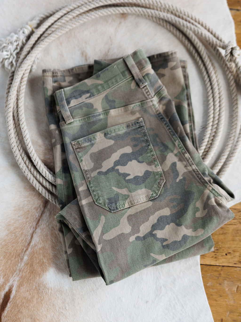 The Granger High Rise Wide Leg Jean by Savannah Sevens western life{&}style, folded in a camo pattern, rests on a light animal hide rug with a coiled rope nearby on a wooden floor.