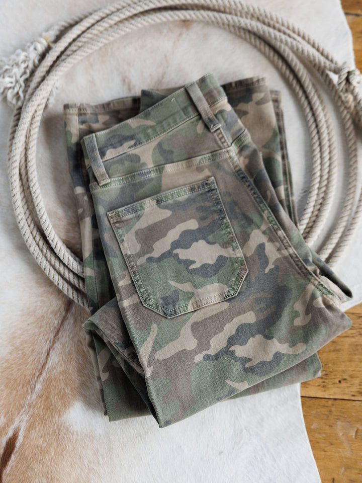 The Granger High Rise Wide Leg Jean by Savannah Sevens western life{&}style, folded in a camo pattern, rests on a light animal hide rug with a coiled rope nearby on a wooden floor.