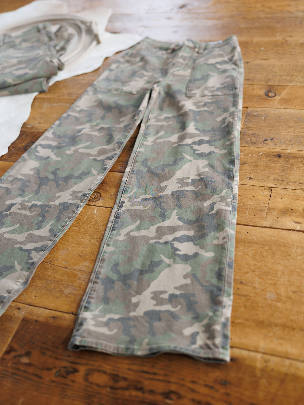 The Granger High Rise Wide Leg Jean by Savannah Sevens western life{&}style in a camo pattern lies flat on a wooden floor beside another folded garment.