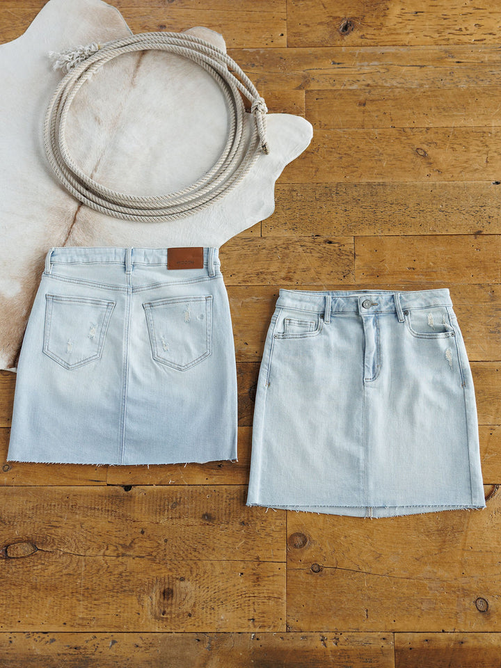 Two The Sumner Skirts by Savannah Sevens western life{&}style are laid flat: one shows the back with pockets and a patch, the other the front with buttons, pockets, and a raw hem. A coiled rope sits on a white animal hide in the corner.