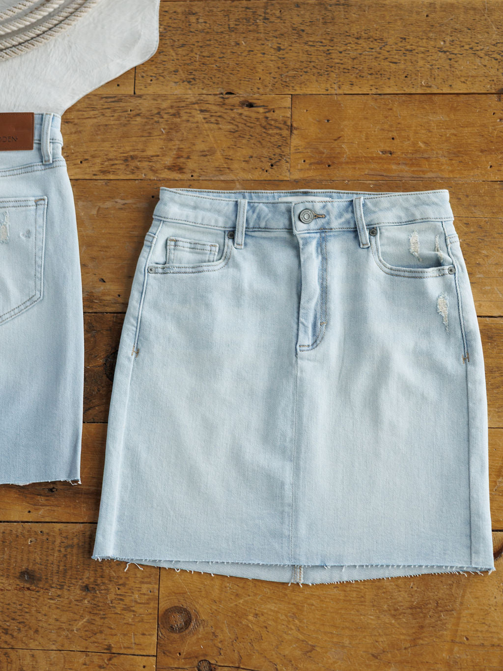 The Sumner Skirt by Savannah Sevens western life{&}style is a light wash denim skirt featuring a raw cut hem and subtle distressing, shown laid flat on wood beside a coordinating denim piece.