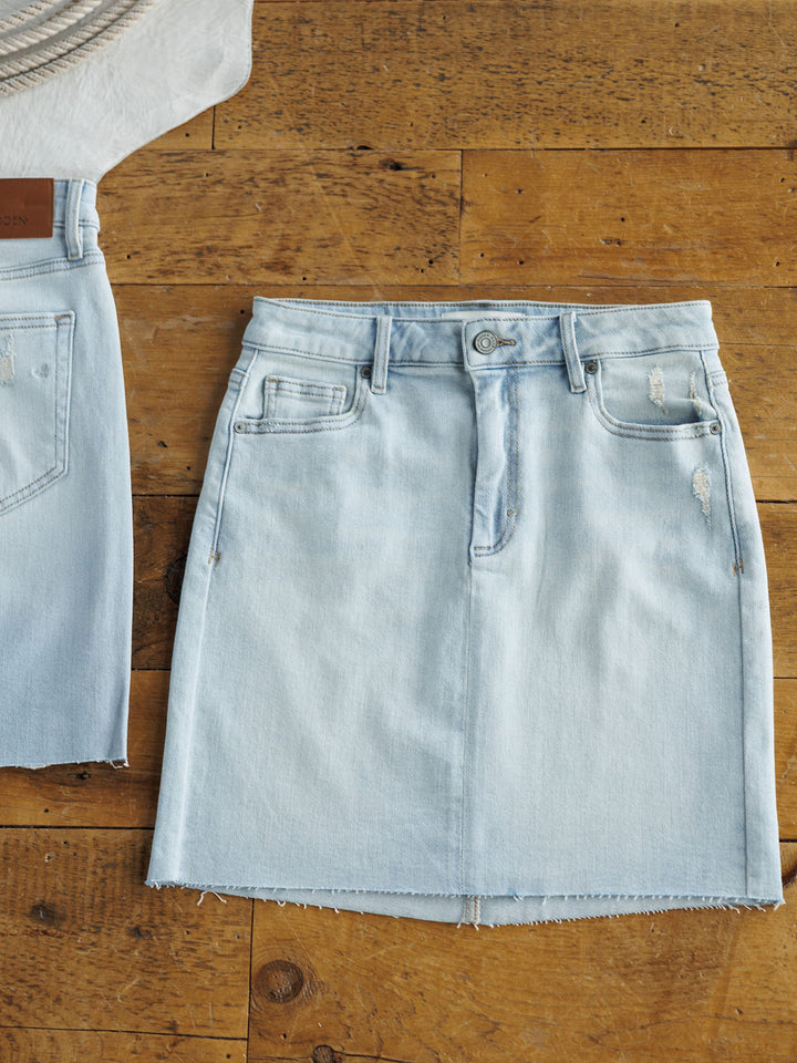 The Sumner Skirt by Savannah Sevens western life{&}style is a light wash denim skirt featuring a raw cut hem and subtle distressing, shown laid flat on wood beside a coordinating denim piece.