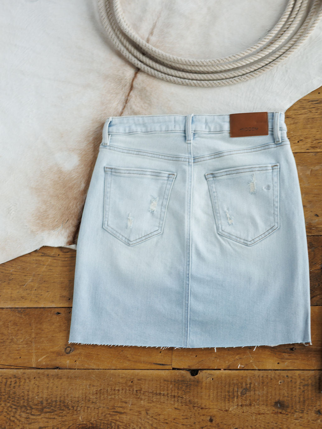 The Sumner Skirt by Savannah Sevens western life{&}style—a light wash distressed denim skirt with back pockets and a raw cut hem—rests on a wooden floor beside a cowhide rug and coiled rope.