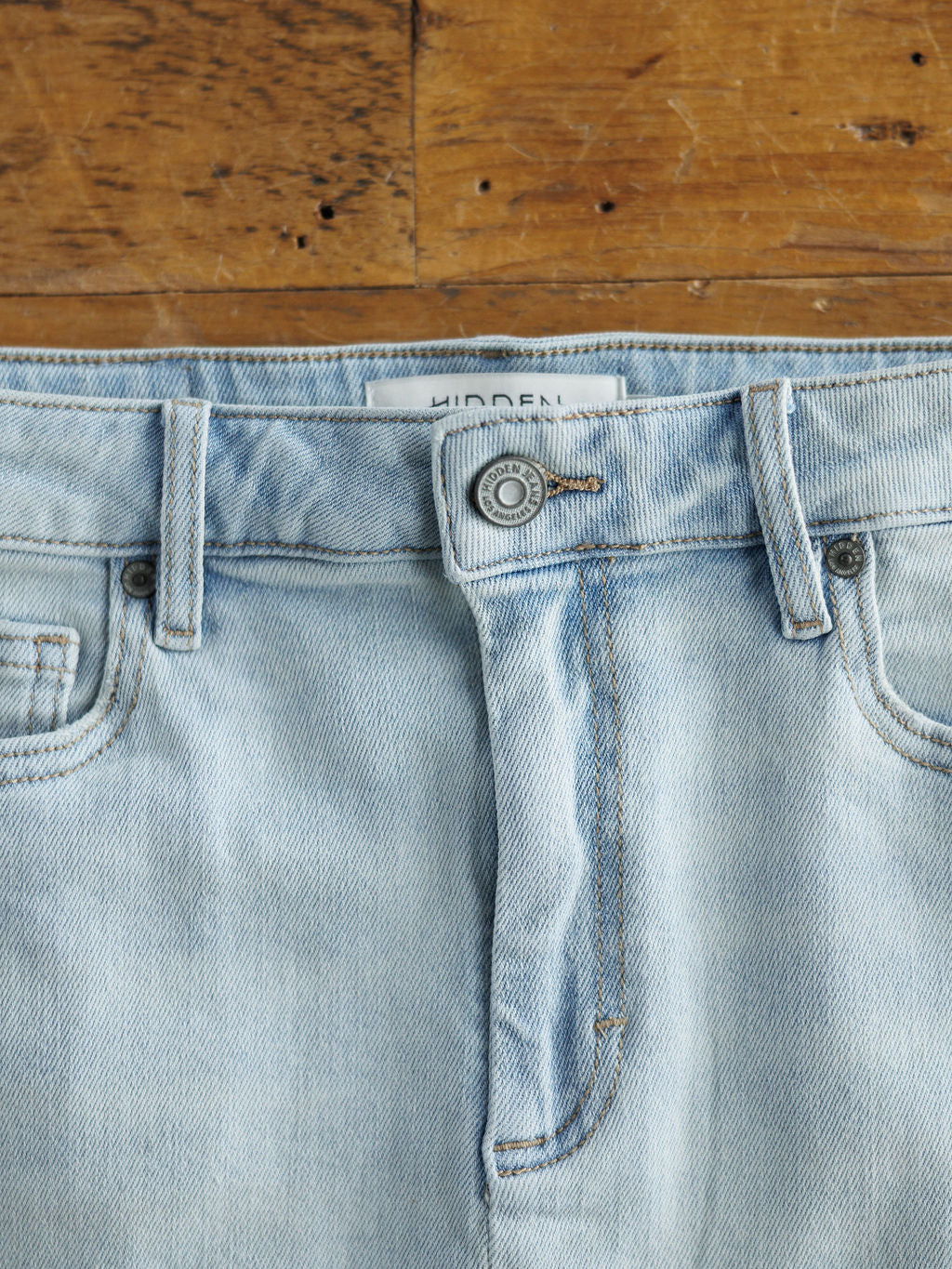 A close-up of The Sumner Skirt by Savannah Sevens western life{&}style shows its light wash denim waistband and silver button fly, set against a wooden background.