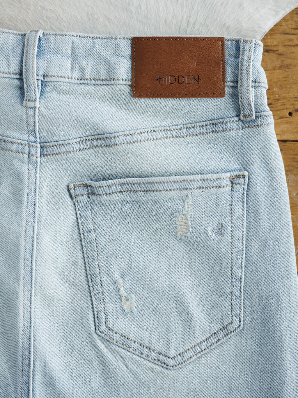 The Sumner Skirt by Savannah Sevens western life{&}style features light wash denim with a raw cut hem, a back pocket with subtle distressing, and a brown leather patch above the pocket. The skirt is displayed on a wooden surface.