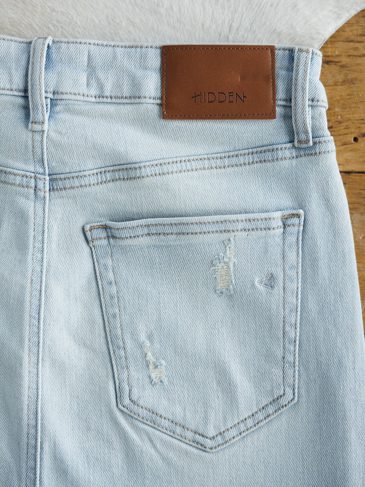 The Sumner Skirt by Savannah Sevens western life{&}style features light wash denim with a raw cut hem, a back pocket with subtle distressing, and a brown leather patch above the pocket. The skirt is displayed on a wooden surface.