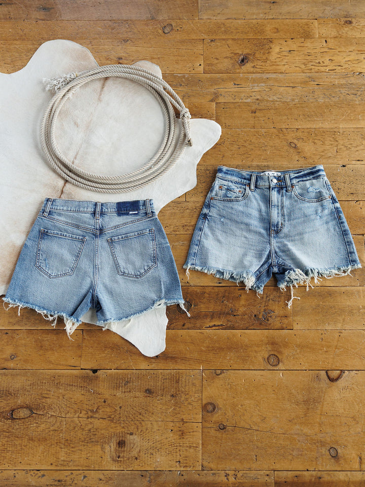 Two pairs of The Paycee Shorts by Savannah Sevens western life{&}style rest on a wooden floor. Both feature a medium light wash and raw cut frayed hems, arranged next to a coiled rope on a white animal hide rug.