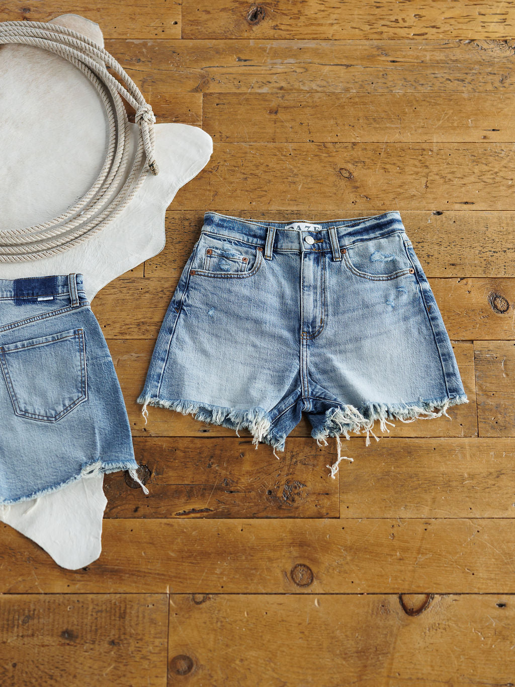 The Paycee Shorts by Savannah Sevens western life{&}style, featuring a high rise and medium light wash with a raw frayed hem, are displayed on a wooden floor beside a coiled rope and another pair of denim shorts.
