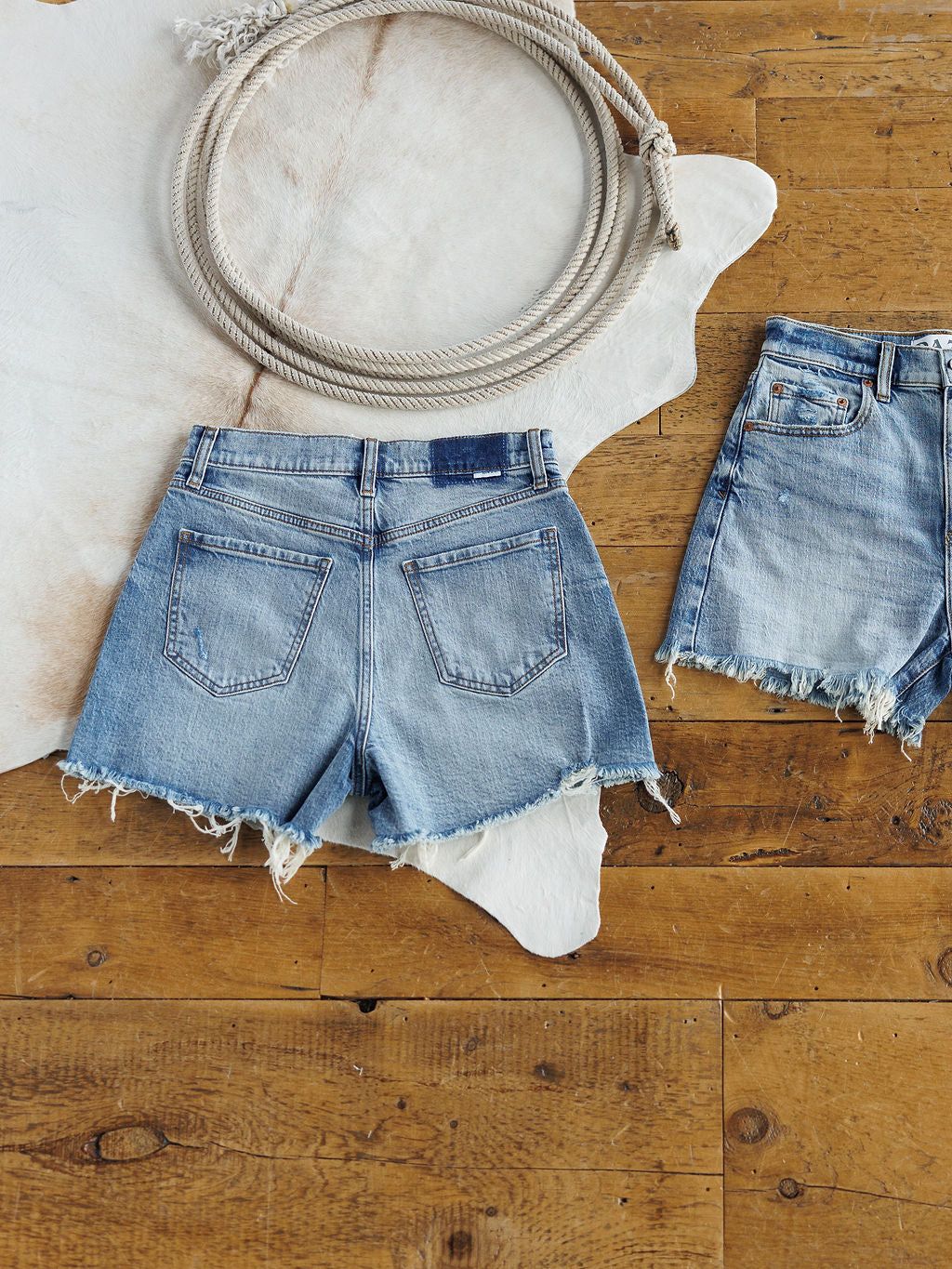 Two pairs of The Paycee Shorts by Savannah Sevens western life{&}style, featuring high rise, medium light wash denim and a raw cut frayed hem, are laid out on a wooden floor next to a cowhide rug.
