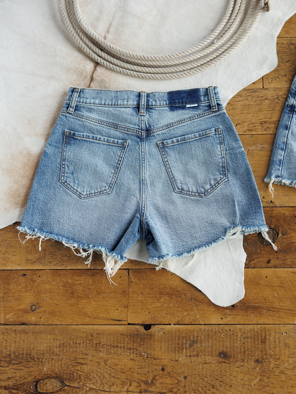 The Paycee Shorts by Savannah Sevens western life{&}style are high rise, medium light wash denim shorts with a raw frayed hem, shown lying flat on wood flooring partly over a white cowhide rug, with a coiled rope at the top of the image.