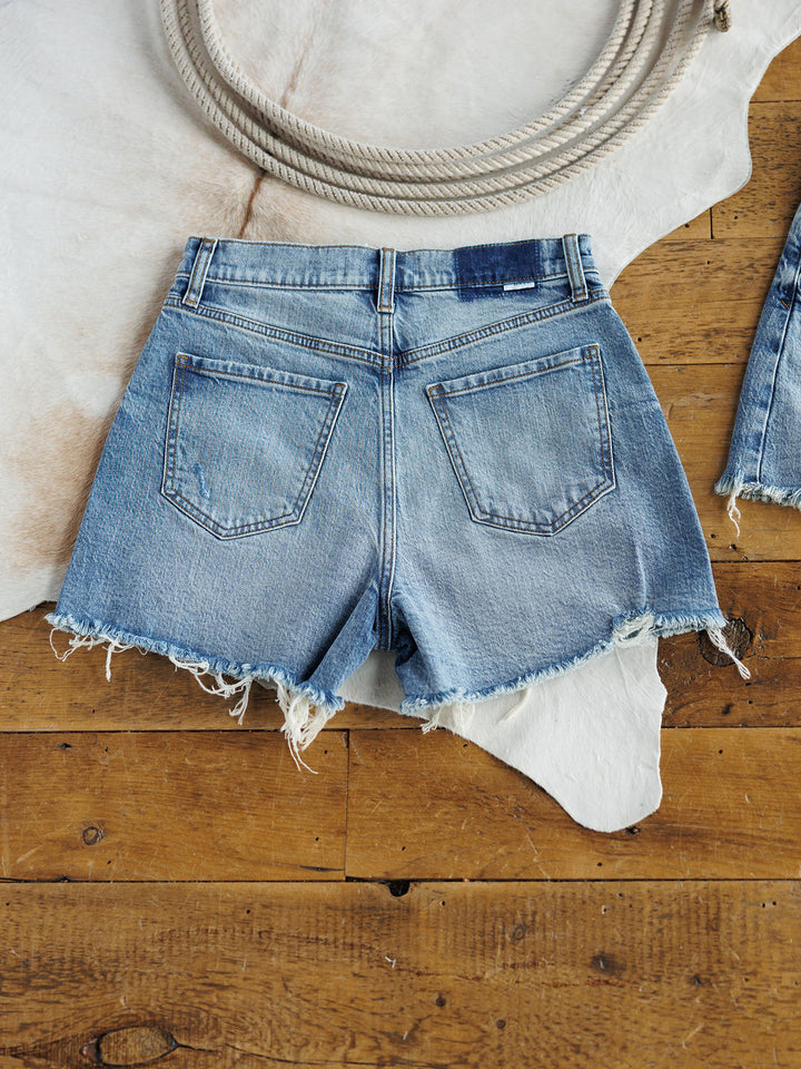 The Paycee Shorts by Savannah Sevens western life{&}style are high rise, medium light wash denim shorts with a raw frayed hem, shown lying flat on wood flooring partly over a white cowhide rug, with a coiled rope at the top of the image.