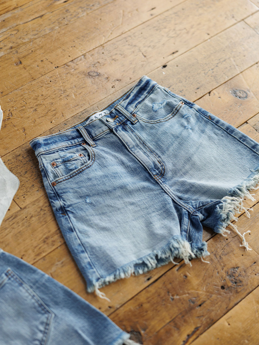 The Paycee Shorts by Savannah Sevens western life{&}style are high rise, medium light wash denim shorts with raw cut frayed hems, front pockets, belt loops, and a button closure, shown on a rustic wooden floor.