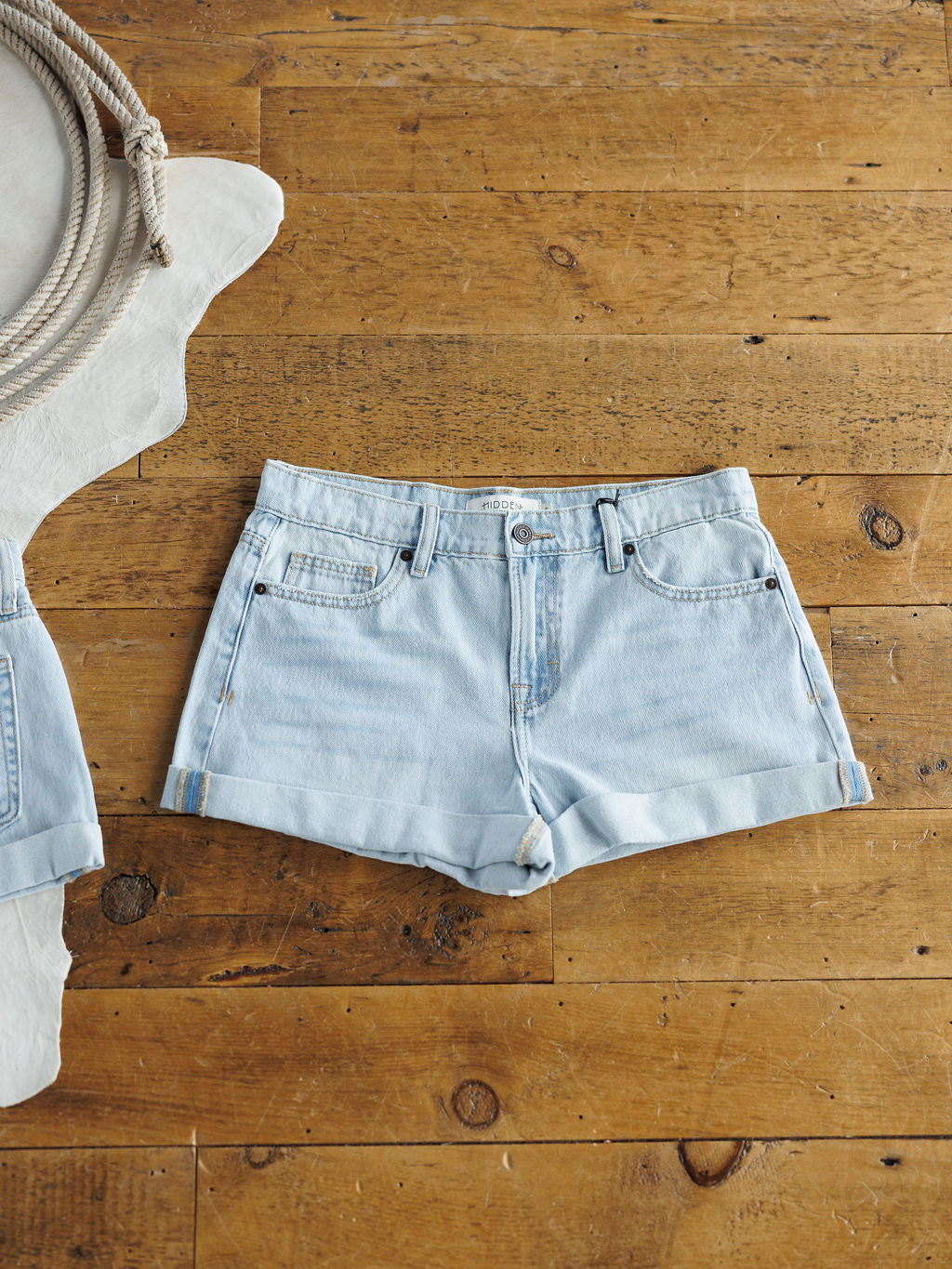 The Mackie Shorts by Savannah Sevens western life{&}style, light wash denim with rolled cuffs, are laid flat on a rustic wooden floor beside a coiled rope and a white animal hide.