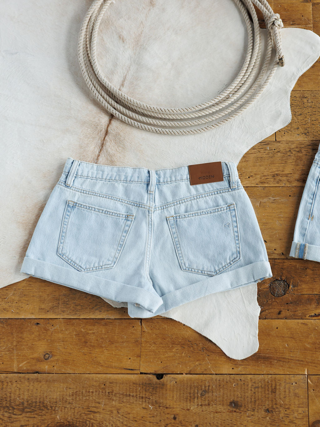 The Mackie Shorts by Savannah Sevens western life{&}style are light wash, high rise denim shorts with two back pockets and a brown leather patch, shown laid flat on a white cowhide rug beside a coiled rope on a wooden floor.
