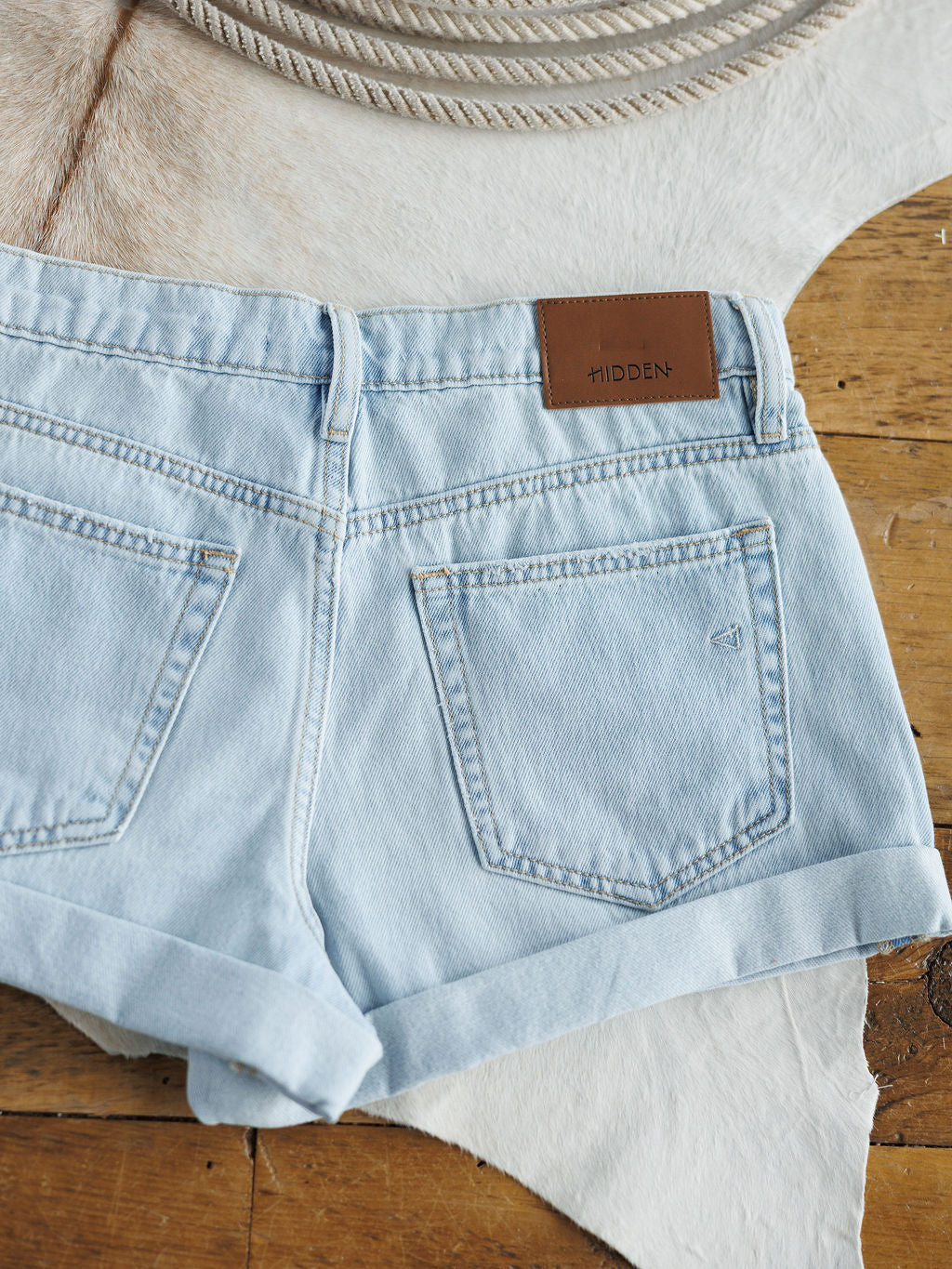 The Mackie Shorts by Savannah Sevens western life{&}style feature light wash denim, rolled cuffs, and a brown leather HIDDEN patch on the waistband, displayed flat on white cowhide and wood with coiled rope in the background.
