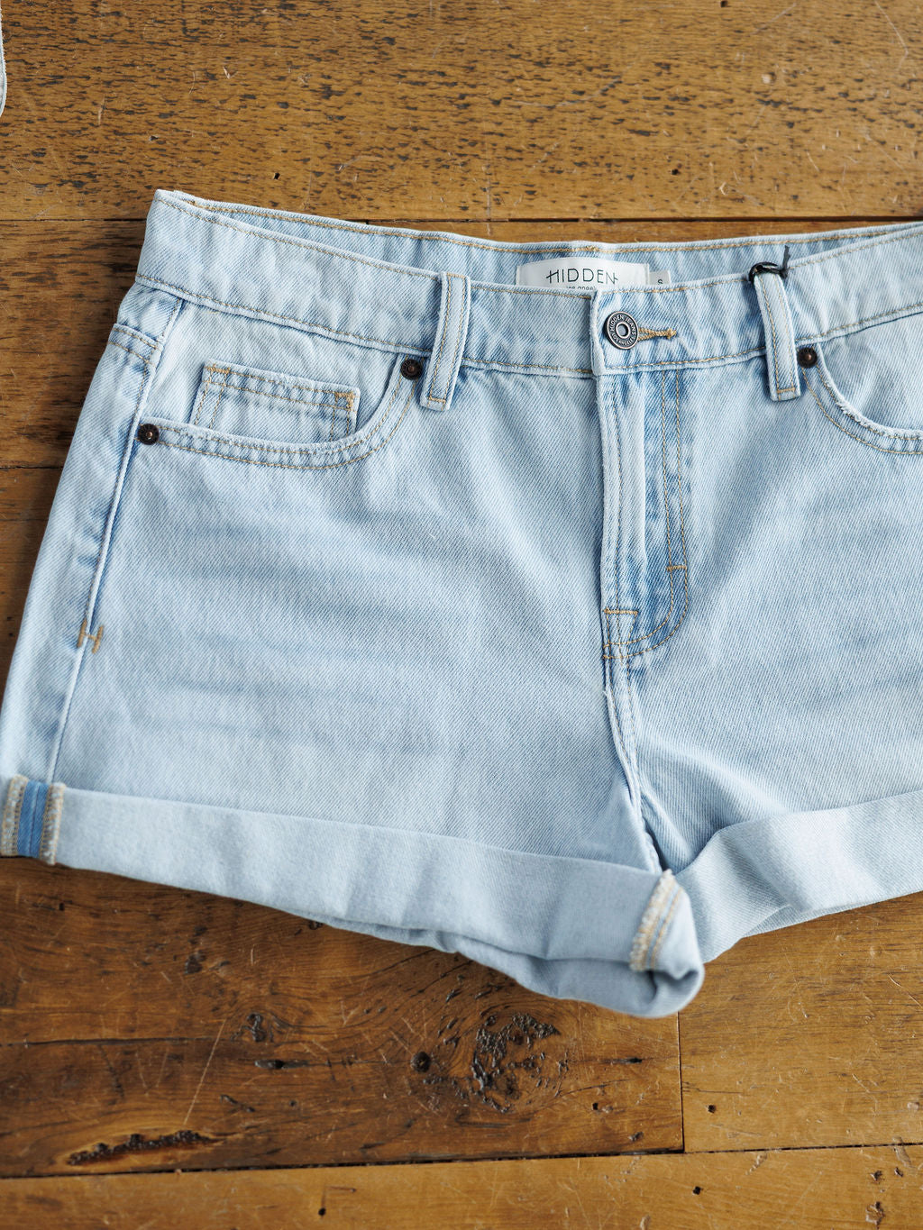 The Mackie Shorts by Savannah Sevens western life{&}style are light wash denim with rolled hems, front and coin pockets, belt loops, and a button-zipper closure, shown laid flat on a rustic wooden surface.